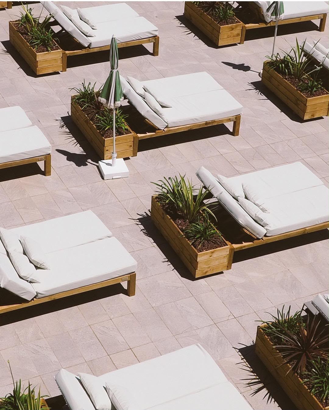 Outdoor patio lounge chairs with white cushions arranged on a light stone tile surface, interspersed with rectangular wooden planters containing green plants. A closed green-and-white patio umbrella stands near the center.