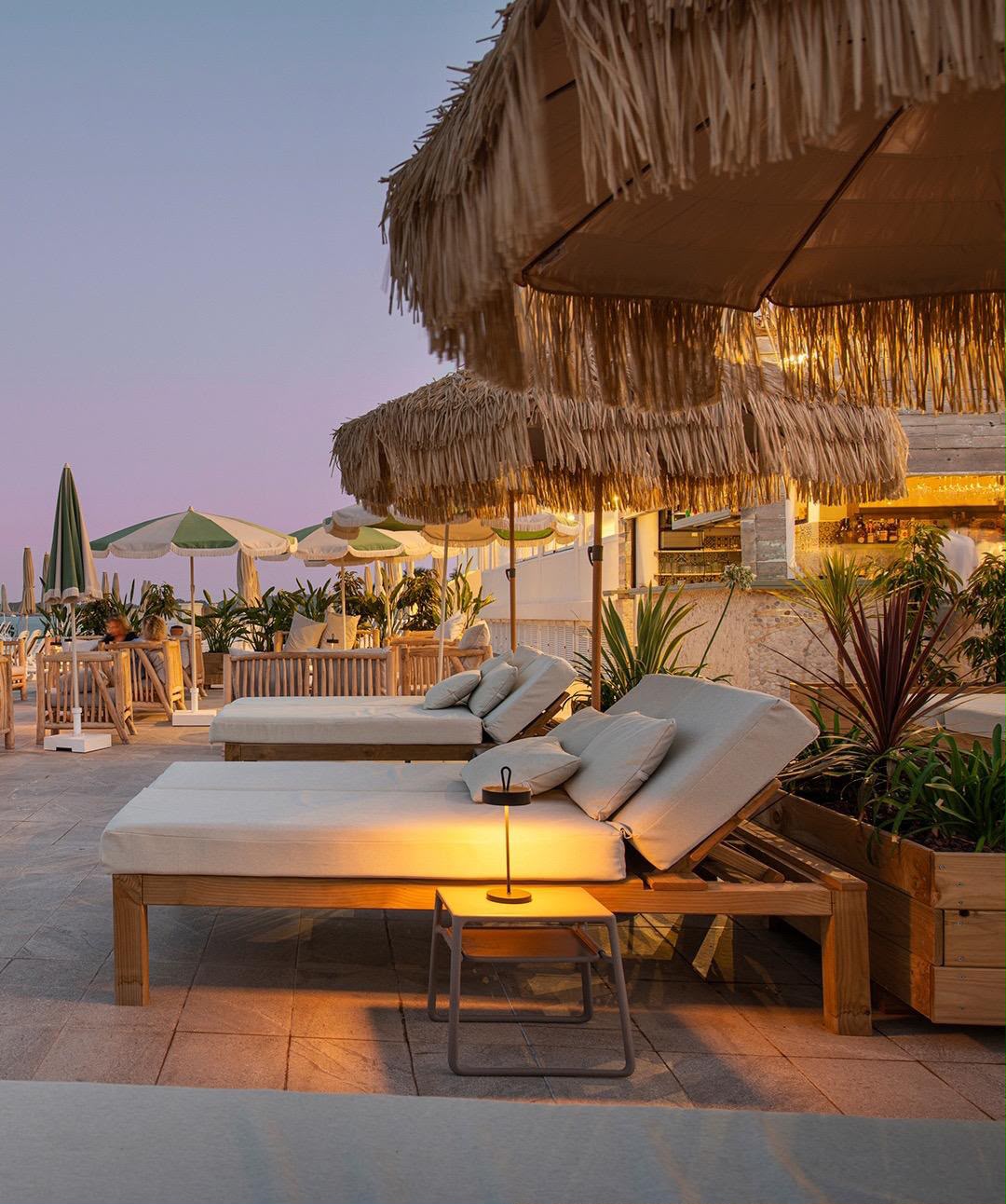 Outdoor beach lounge setup at dusk with wooden chaise lounges, thatched umbrellas, small side tables with warm lights, and tropical plants in a patio setting.