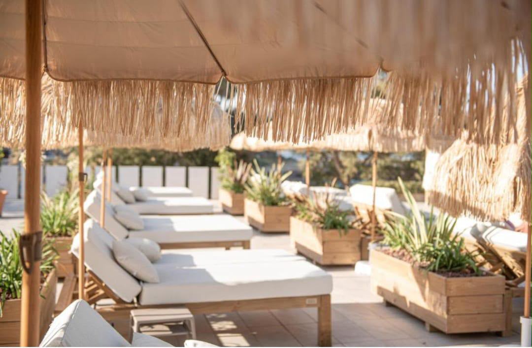 Outdoor patio seating area under a large straw-fringed umbrella, featuring multiple white cushioned lounge chairs, wooden planters with greenery, and a warm neutral color palette.