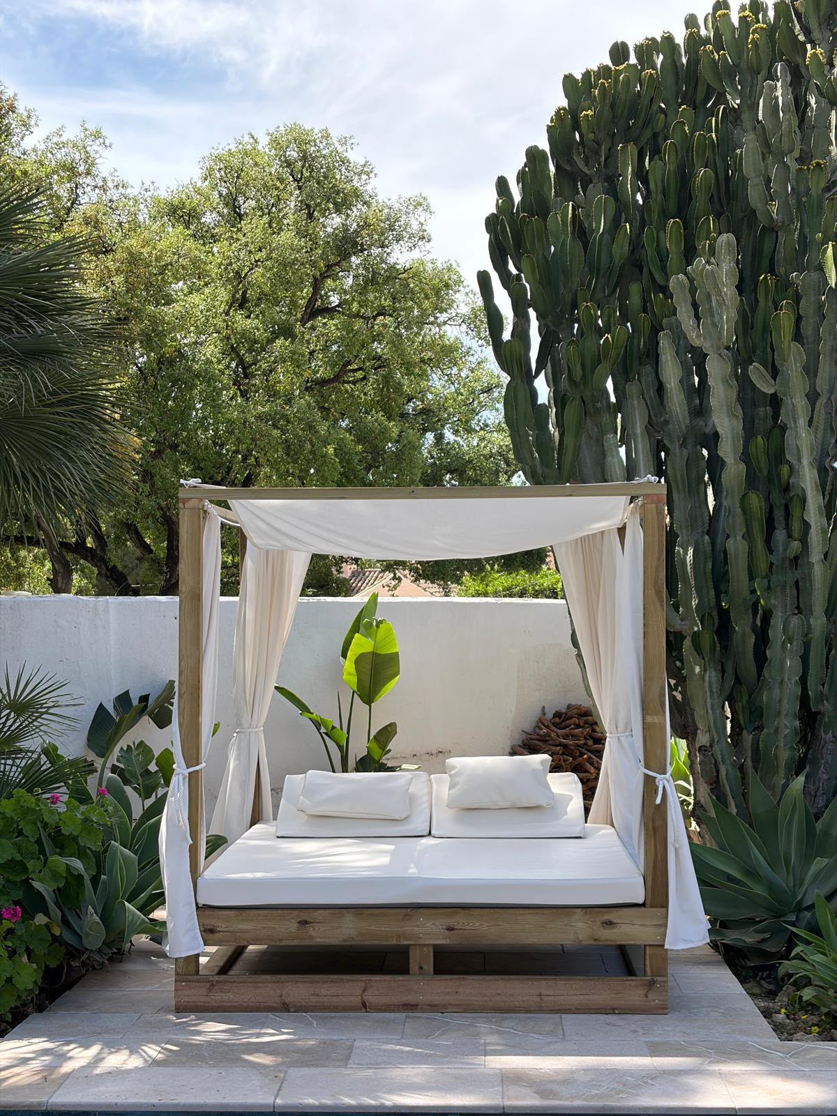 A wooden outdoor daybed with a canopy and draped white fabric, styled with white cushions and pillows, set on a stone patio in a lush garden with cacti and trees.