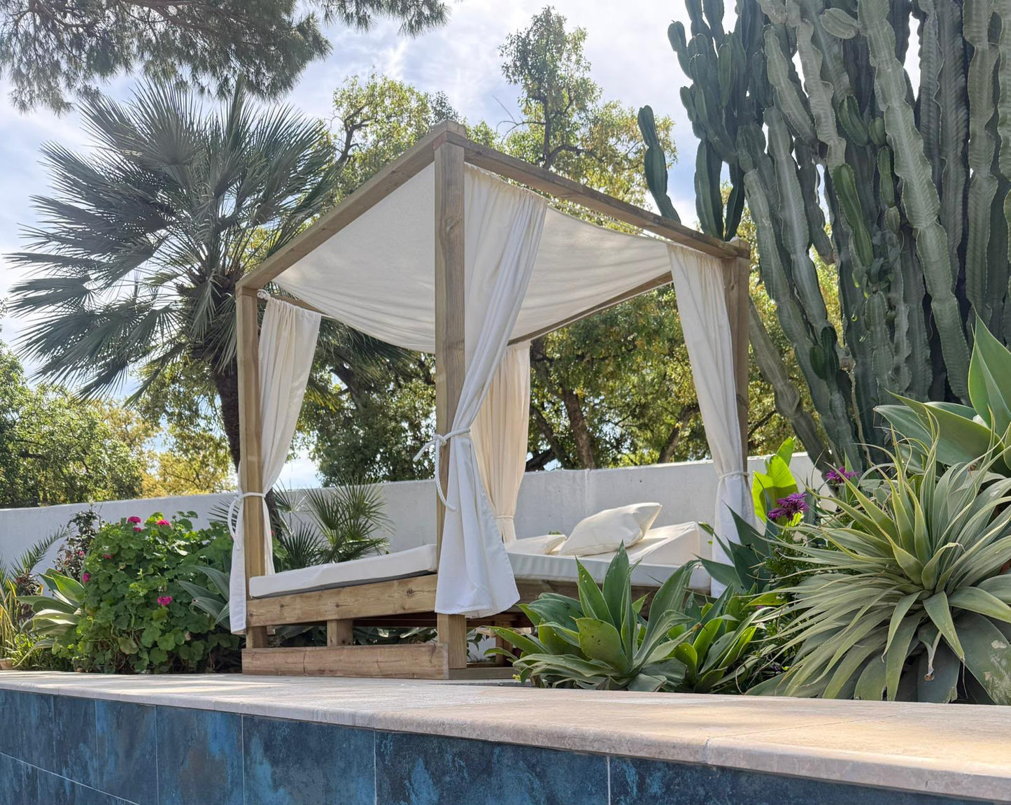 A wooden four-poster canopy daybed with draped off-white fabric curtains, set outdoors in a garden next to a swimming pool with cacti and palm trees in the background.
