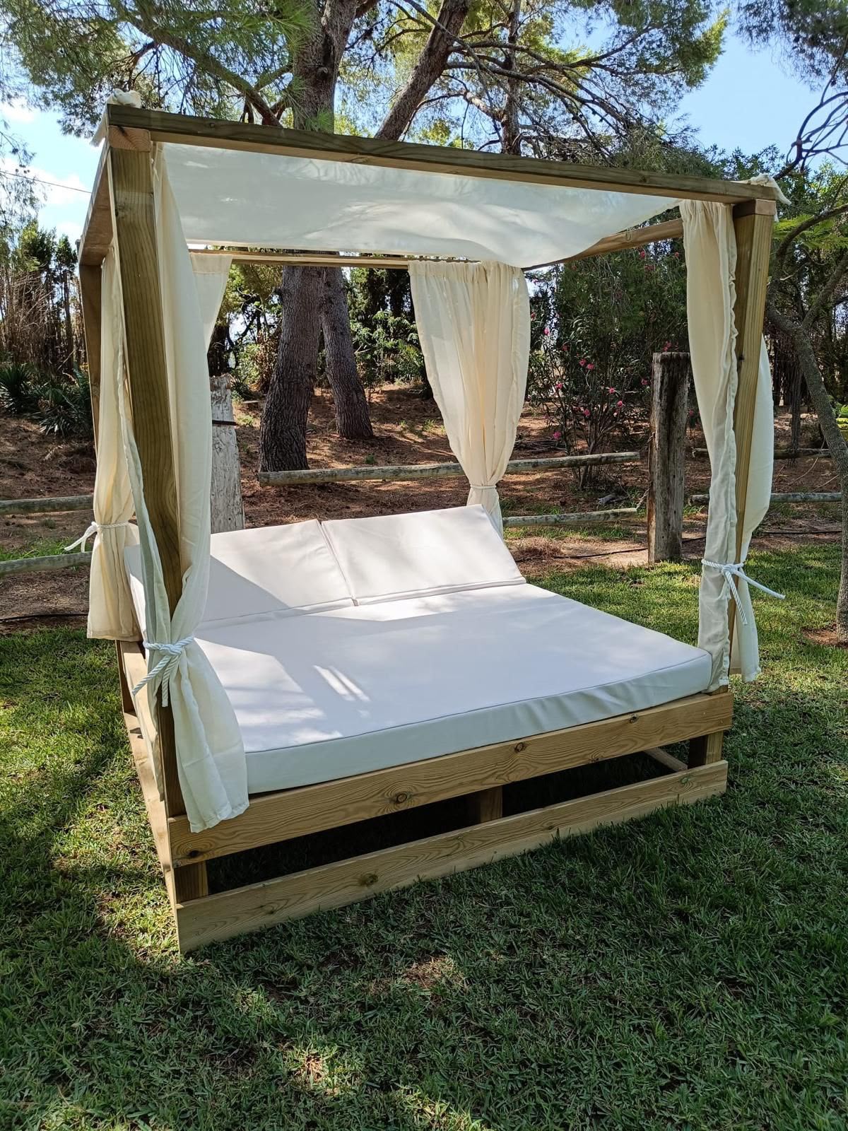 A wooden four-poster daybed with a canopy and cream-colored curtains, set outdoors on a grassy lawn with trees in the background.